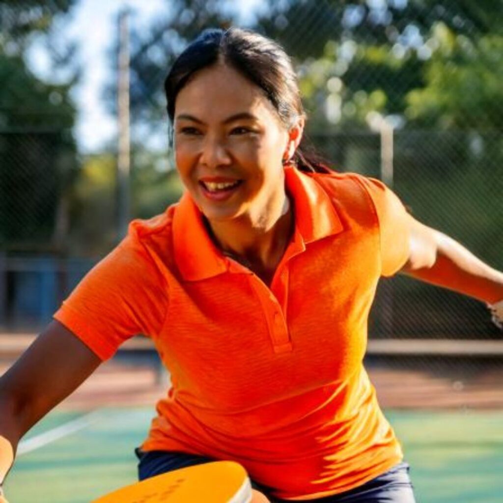 Woman playing pickleball outdoors in orange shirt.